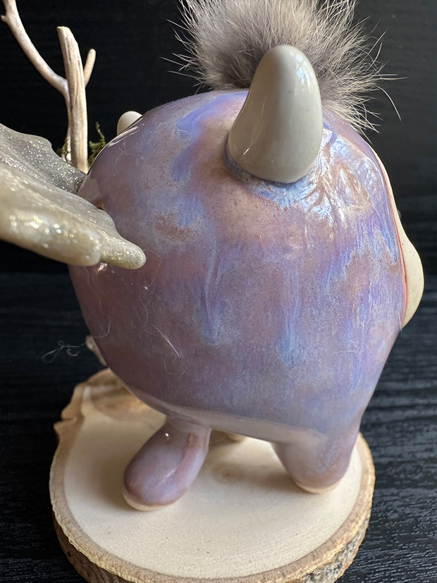 Ceramic sculpture of a stylized cute troll monster, with a large face and rounded body with feet and no arms. It has a fluff atop its head and stands on a wooden base with a branch behind it and crystals.