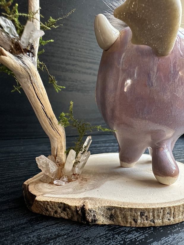 Ceramic sculpture of a stylized cute troll monster, with a large face and rounded body with feet and no arms. It has a fluff atop its head and stands on a wooden base with a branch behind it and crystals.