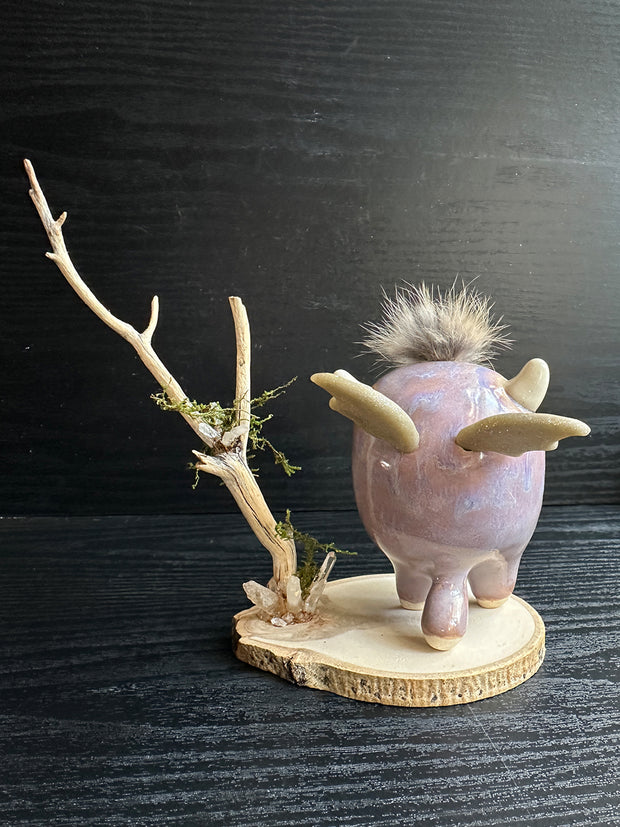 Ceramic sculpture of a stylized cute troll monster, with a large face and rounded body with feet and no arms. It has a fluff atop its head and stands on a wooden base with a branch behind it and crystals.