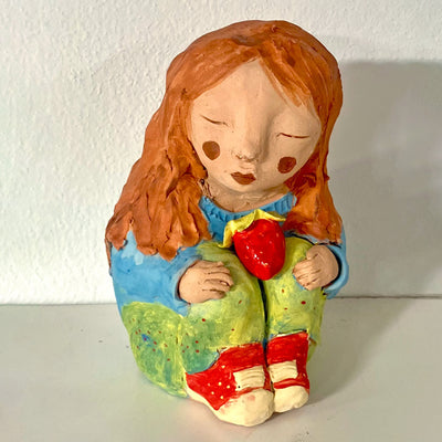 Sculpture of a orange haired girl sitting and hugging her knees to her chest. She wears a blue sweater, green pants and red sneakers. A strawberry rests atop her knees.