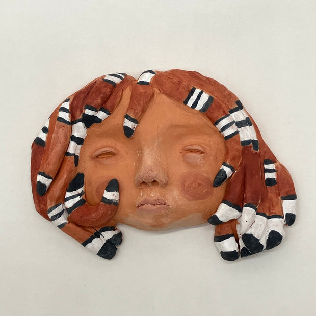 Terracotta sculpture of a person's face, with the back flat so it lays flat. Their hair is comprised of snakes, dark orange with black and white striping.