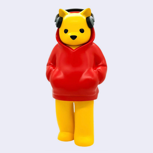 Vinyl figure of with a yellow bear with a humanlike body, standing with his hands in the pockets of a red hoodie. It wears a pair of headphones atop its ears.