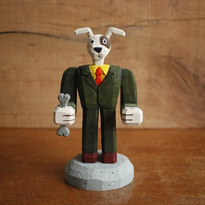 Painted whittled wooden sculpture of a dog with large floppy ears and a humanoid body, dressed as a business man holding a bone in one hand.