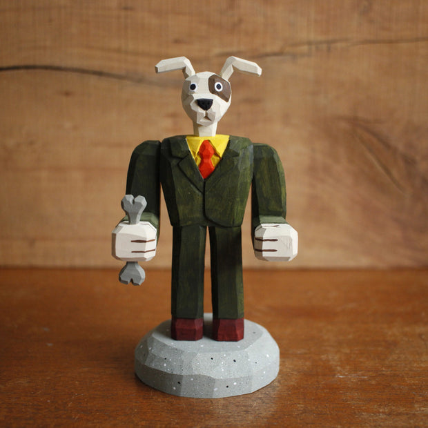 Painted whittled wooden sculpture of a dog with large floppy ears and a humanoid body, dressed as a business man holding a bone in one hand.