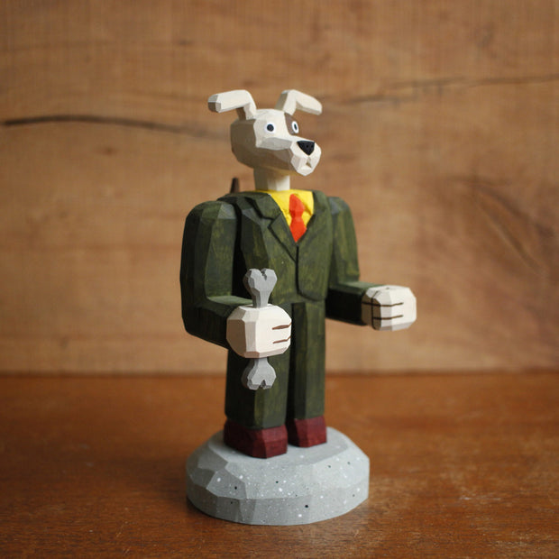 Painted whittled wooden sculpture of a dog with large floppy ears and a humanoid body, dressed as a business man holding a bone in one hand.