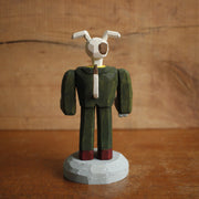 Painted whittled wooden sculpture of a dog with large floppy ears and a humanoid body, dressed as a business man holding a bone in one hand.