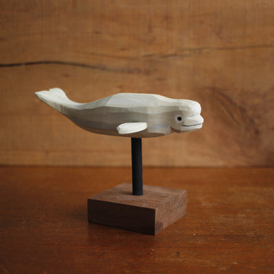 Painted whittled wooden sculpture of a white beluga whale, with a subtle smile. It hovers above a small wooden block stand by a black thin rod.