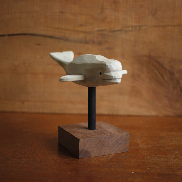 Painted whittled wooden sculpture of a white beluga whale, with a subtle smile. It hovers above a small wooden block stand by a black thin rod.