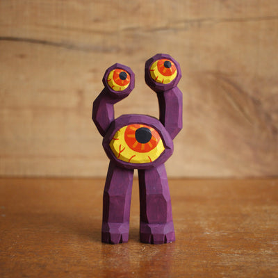 Painted whittled wooden sculpture of a purple 3 eyed monster with 2 legs, one of the eyes taking up the majority of the body and the other 2 acting as appendages. 