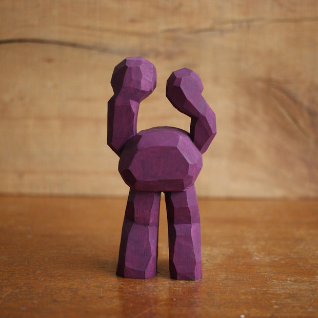 Painted whittled wooden sculpture of a purple 3 eyed monster with 2 legs, one of the eyes taking up the majority of the body and the other 2 acting as appendages. 