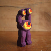 Painted whittled wooden sculpture of a purple 3 eyed monster with 2 legs, one of the eyes taking up the majority of the body and the other 2 acting as appendages. 