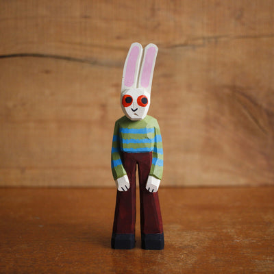 Painted whittled wooden sculpture of a white rabbit with red eyes and a humanoid body, wearing a green and blue striped sweater and brown pants.