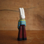 Painted whittled wooden sculpture of a white rabbit with red eyes and a humanoid body, wearing a green and blue striped sweater and brown pants.