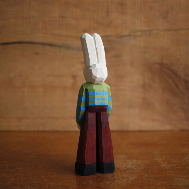 Painted whittled wooden sculpture of a white rabbit with red eyes and a humanoid body, wearing a green and blue striped sweater and brown pants.