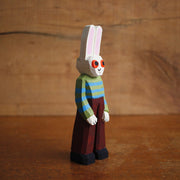 Painted whittled wooden sculpture of a white rabbit with red eyes and a humanoid body, wearing a green and blue striped sweater and brown pants.