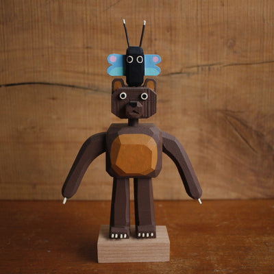 Assembled painted whittled wooden sculpture of a bear with long arms, standing on its hind legs with a small black and blue butterfly atop its head.