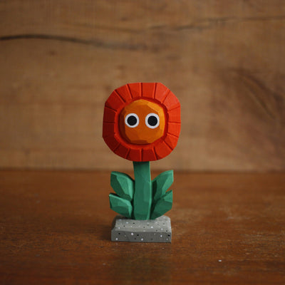 Small painted whittled wooden sculpture of a red and orange flower with a green stem and a pair of simple, cute cartoon eyes. Its positioned on a stone colored tile.