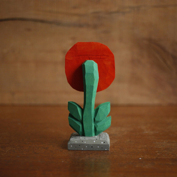 Small painted whittled wooden sculpture of a red and orange flower with a green stem and a pair of simple, cute cartoon eyes. Its positioned on a stone colored tile.