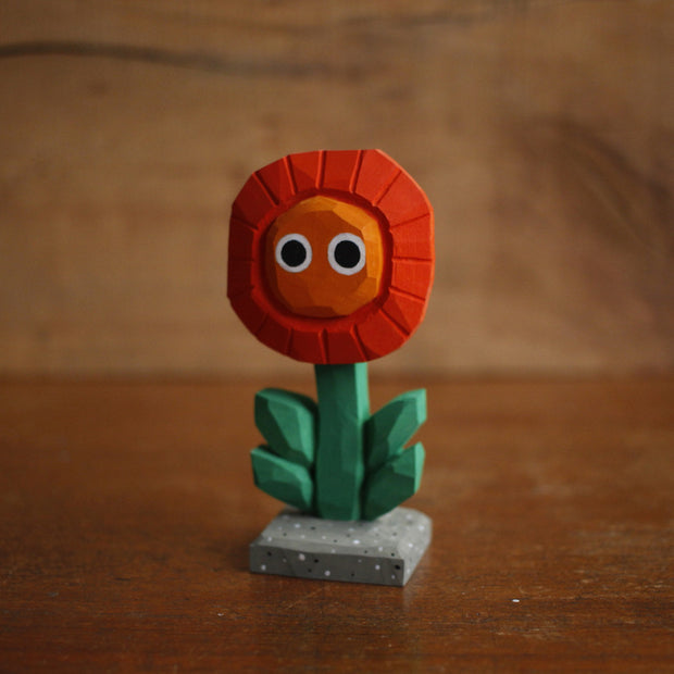 Small painted whittled wooden sculpture of a red and orange flower with a green stem and a pair of simple, cute cartoon eyes. Its positioned on a stone colored tile.