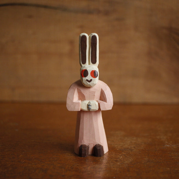 Painted whittled wooden sculpture of a small white bunny with very tall ears and red eyes. It has a humanoid body, wearing a pink dress and looking off to the side with its hands folded.