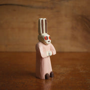 Painted whittled wooden sculpture of a small white bunny with very tall ears and red eyes. It has a humanoid body, wearing a pink dress and looking off to the side with its hands folded.