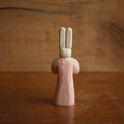 Painted whittled wooden sculpture of a small white bunny with very tall ears and red eyes. It has a humanoid body, wearing a pink dress and looking off to the side with its hands folded.