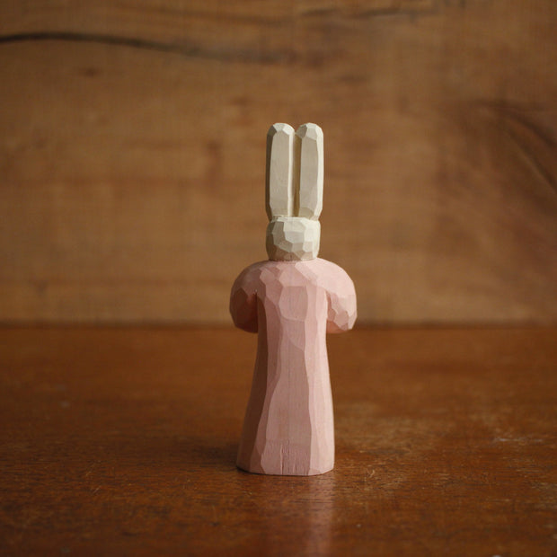 Painted whittled wooden sculpture of a small white bunny with very tall ears and red eyes. It has a humanoid body, wearing a pink dress and looking off to the side with its hands folded.