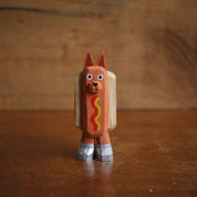 Small painted whittled wood sculpture of a dog dressed as a hot dog, with strips of mustard and ketchup. It wears a pair of silver shoes.