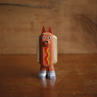 Small painted whittled wood sculpture of a dog dressed as a hot dog, with strips of mustard and ketchup. It wears a pair of silver shoes.