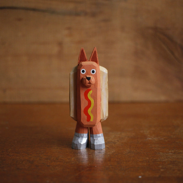 Small painted whittled wood sculpture of a dog dressed as a hot dog, with strips of mustard and ketchup. It wears a pair of silver shoes.