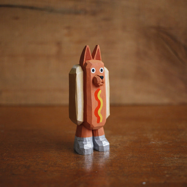 Small painted whittled wood sculpture of a dog dressed as a hot dog, with strips of mustard and ketchup. It wears a pair of silver shoes.