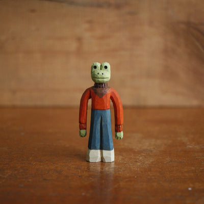 Small painted whittled wood sculpture of a frog with a humanoid body, skinny and wearing a dark orange sweater and blue jeans.