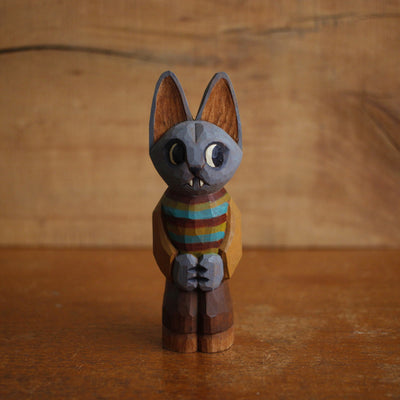 Painted whittled wooden sculpture of a gray cat with a cute expression and pointed teeth. It looks off to the side and has a humanoid body, wearing a striped earth toned sweater and brown pants.