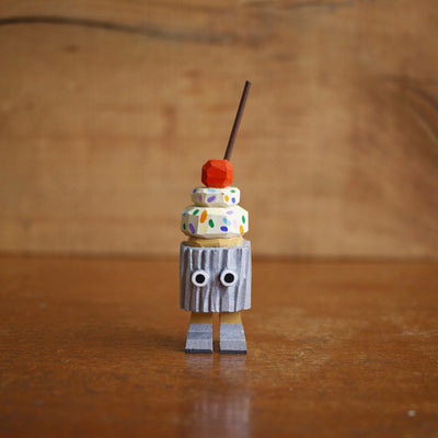 Wooden sculpture of a cupcake with white frosting, sprinkles, and a cherry on top. Its wrapped in a silver wrapper and stands on 2 legs, with a pair of eyes as its only facial feature.