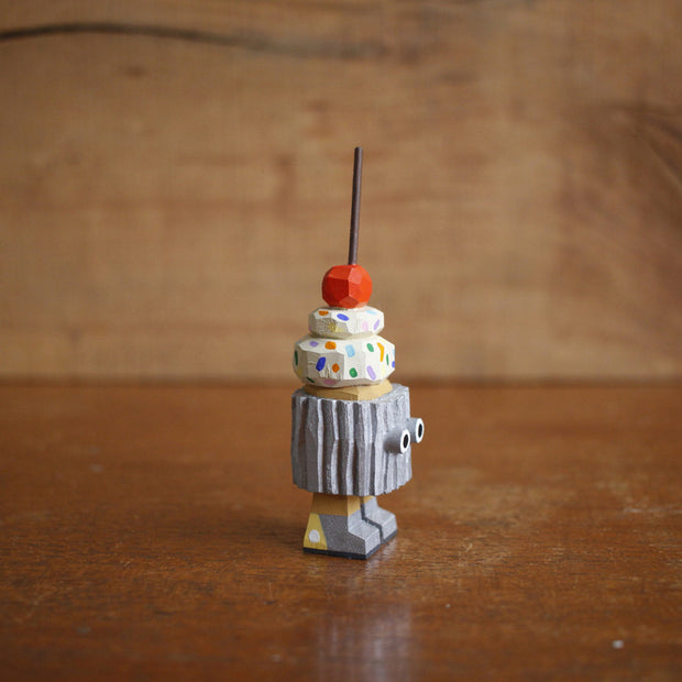 Wooden sculpture of a cupcake with white frosting, sprinkles, and a cherry on top. Its wrapped in a silver wrapper and stands on 2 legs, with a pair of eyes as its only facial feature.