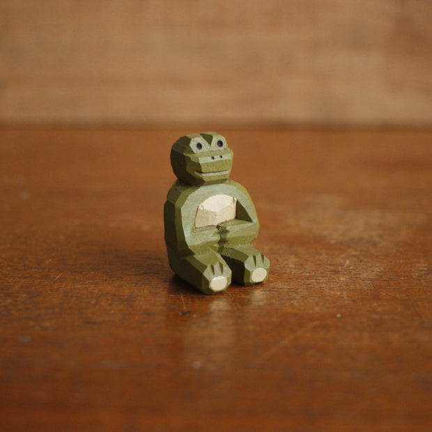 Small painted whittled wood sculpture of a green frog, sitting on the ground with its legs extended out in front and its hands over its stomach.