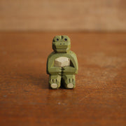 Small painted whittled wood sculpture of a green frog, sitting on the ground with its legs extended out in front and its hands over its stomach.