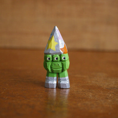 Small whittled wooden sculpture of a 3 eyed green short monster, wearing a silver wizard hat with multicolored stars. It stands on 2 legs, no arms, and wears a pair of silver shoes.