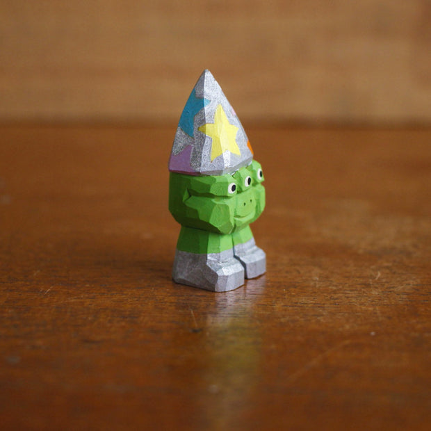 Small whittled wooden sculpture of a 3 eyed green short monster, wearing a silver wizard hat with multicolored stars. It stands on 2 legs, no arms, and wears a pair of silver shoes.