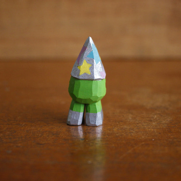 Small whittled wooden sculpture of a 3 eyed green short monster, wearing a silver wizard hat with multicolored stars. It stands on 2 legs, no arms, and wears a pair of silver shoes.