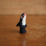 Small painted whittled wooden sculpture of an orca whale, standing on 2 legs with arms that are folded. It has a large sharp toothed open mouth and white eyes.