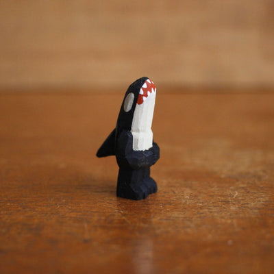 Small painted whittled wooden sculpture of an orca whale, standing on 2 legs with arms that are folded. It has a large sharp toothed open mouth and white eyes.