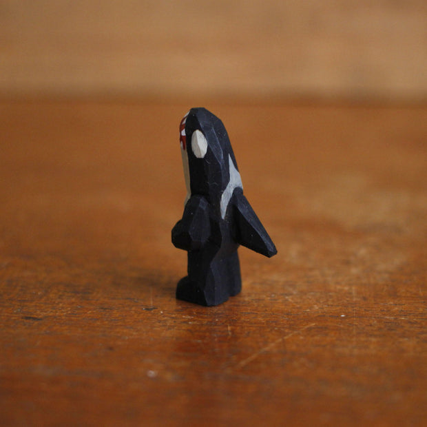 Small painted whittled wooden sculpture of an orca whale, standing on 2 legs with arms that are folded. It has a large sharp toothed open mouth and white eyes.