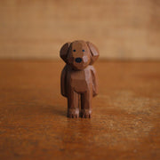 Small painted whittled wooden sculpture of a cute brown dog with floppy ears, standing like a person. It has a set of very small black eyes and a large black nose.