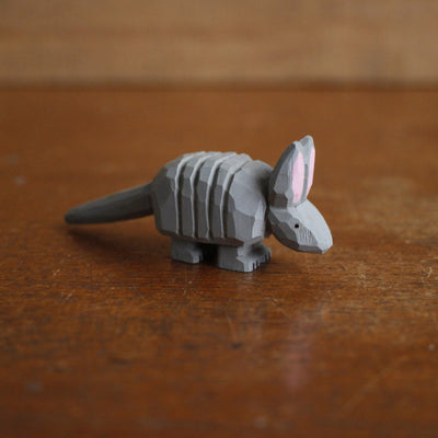 Small whittled wooden sculpture, painted gray, of an armadillo with a rigid back and long tail. It has a simple pair of small black eyes and tall pointed ears.