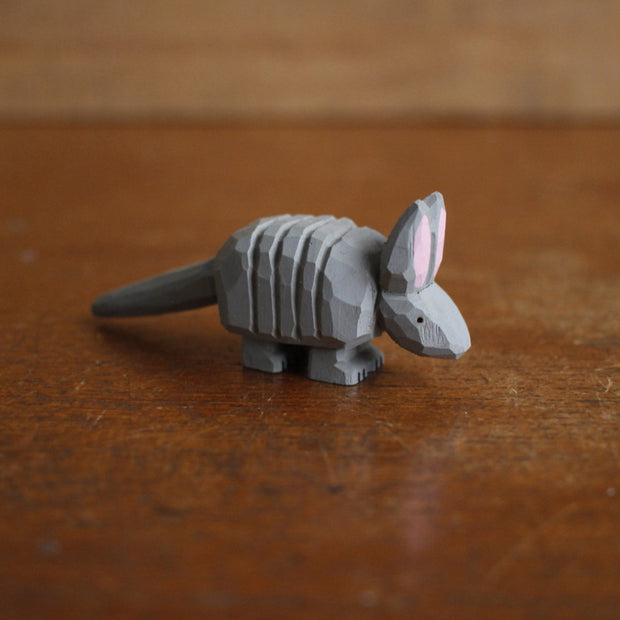 Small whittled wooden sculpture, painted gray, of an armadillo with a rigid back and long tail. It has a simple pair of small black eyes and tall pointed ears.