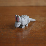 Small whittled wooden sculpture, painted gray, of an armadillo with a rigid back and long tail. It has a simple pair of small black eyes and tall pointed ears.