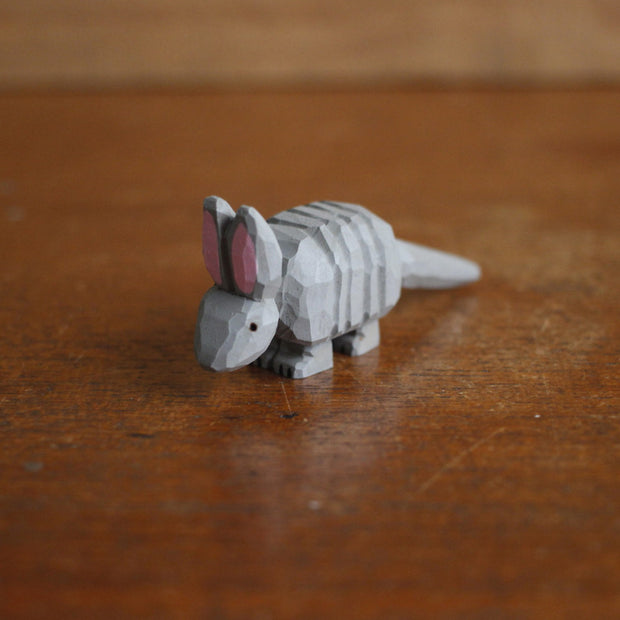 Small whittled wooden sculpture, painted gray, of an armadillo with a rigid back and long tail. It has a simple pair of small black eyes and tall pointed ears.