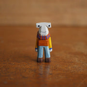 Small painted whittled wooden sculpture of a hammerhead shark, standing on 2 legs like a human and wearing a striped scarf, orange sweater, and blue jeans. It smiles and looks off to the side.