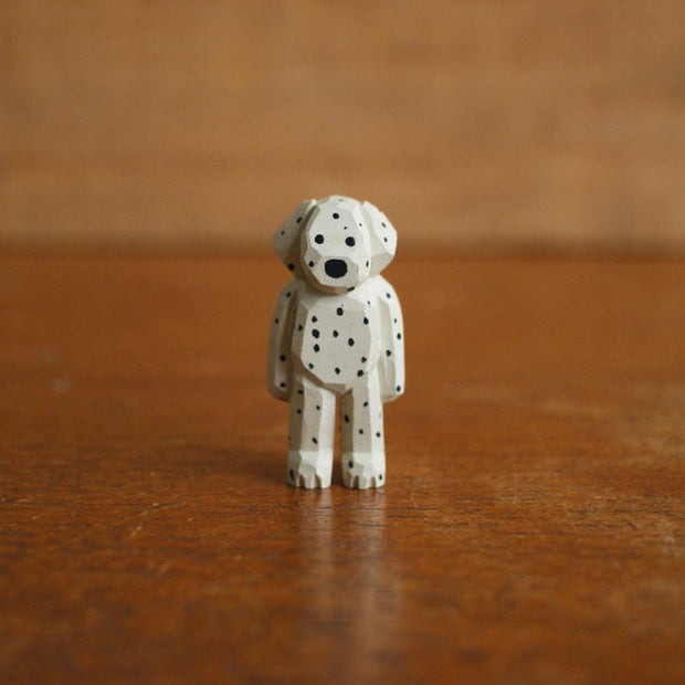 Tiny painted whittled wooden sculpture of a dog with floppy ears, standing on its hind legs like a human. Its white with many black spots, and a small pair of black eyes and a large nose.